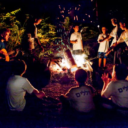 A group of children gathered around a fire, roasting marshmallow at Jewish summer camp.