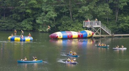 Wide action packed picture of the lake at Camp Ramah Darom with campers in canoes and kayaks, on the water trampoline, swimming and jumping on the blob. There is a high dock with two staff life guards looking on.