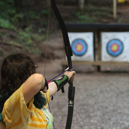 Picture of girl from behind at the Ramah Darom archery range pulling back on a bow and shoot towards a target