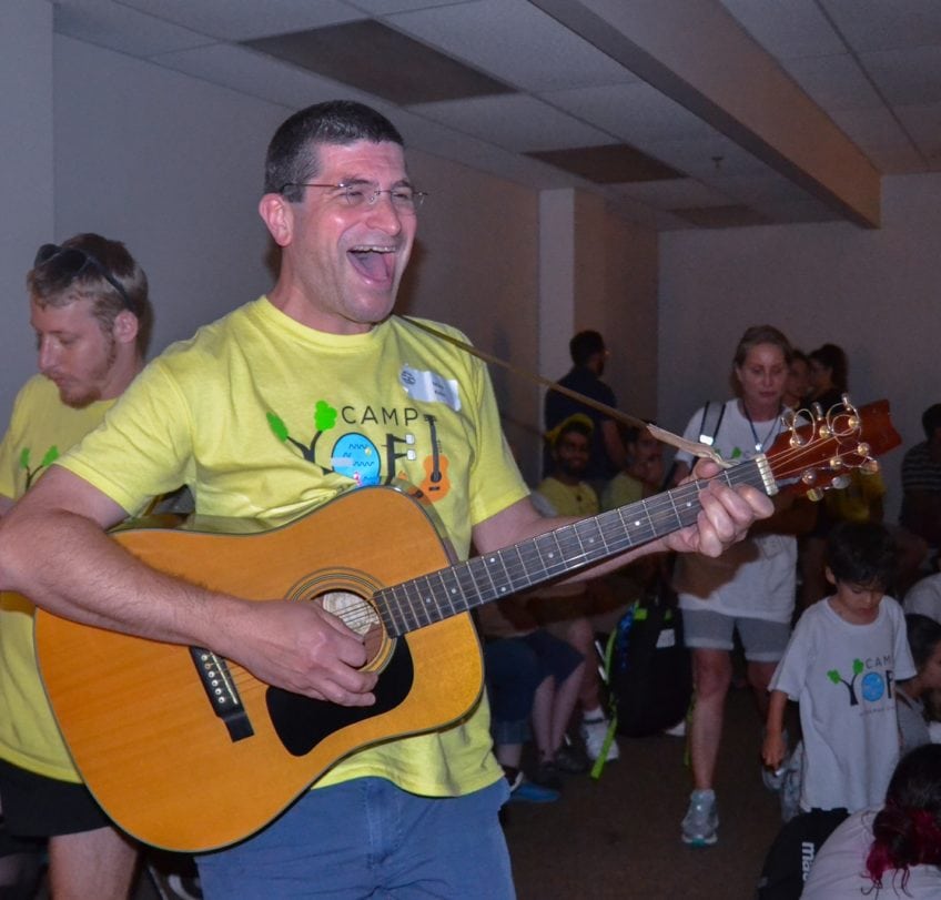 Matt Kalin singing at Camp Yofi with a yellow shirt, a wide smiling mouth and a guitar