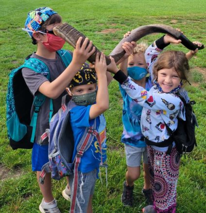 Four young children standing on the kikar, a grassy field at Ramah Darom holding up a large shofar. The three boys are wearing kippot and masks and the girl has a mask under her chin and a huge smile on her face.