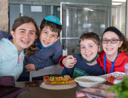 Four children at a table with Passover artwork in front of them. All have big smiles. The girl on the left braces on her teeth. The next boy has Ramah Darom kippah on. The girl on the far right is wearing glasses and has her arm around a younger boy to her left.