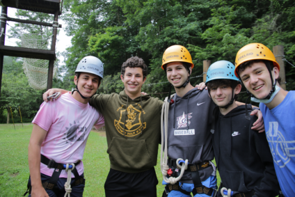 Five teen boys under the high ropes course in protective hats. They have their arms around each other and have big smiles on their faces.